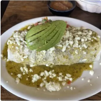 A burrito smothered in green salsa and crumbled queso fresco, topped with sliced avocado.